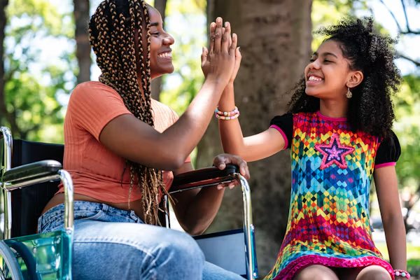 A smiling woman using a wheelchair high-fiving a laughing girl in a park