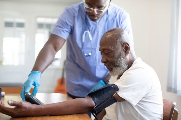 A nurse in blue scrubs checking an older man’s blood pressure at a table