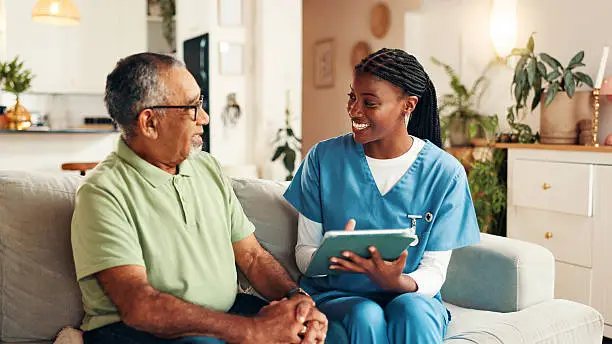 A smiling caregiver in blue scrubs showing a tablet to an older man on a sofa at home